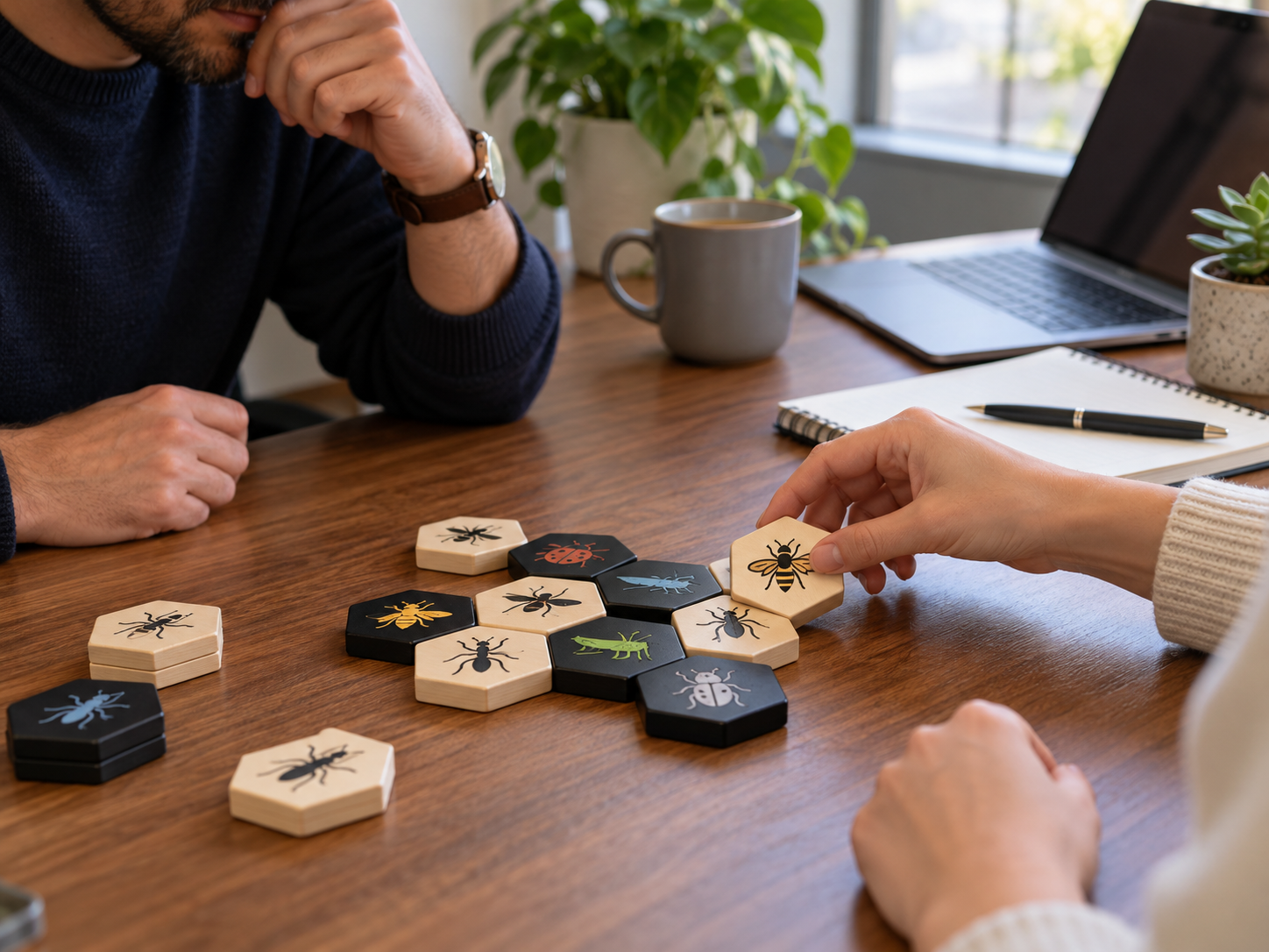 Two adults playing an insect-themed hex-tile strategy game at an office desk